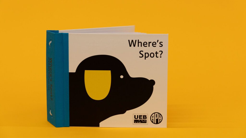 Where's Spot? storybook placed upright on a yellow background. On the cover is the title and a black dog with a yellow ear with the UEB and APH logos in the bottom right corner.
