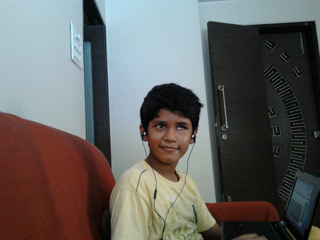 A young Bhavya, wearing a yellow tshirt and sitting on an orange couch, smiles at the camera. He is wearing wired black headphones.