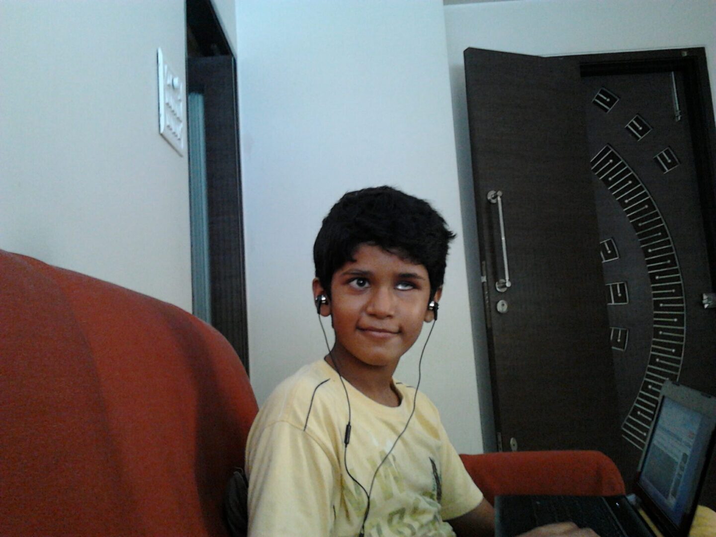 A young Bhavya, wearing a yellow tshirt and sitting on an orange couch, smiles at the camera. He is wearing wired black headphones.