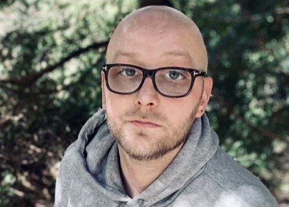 Alex, wearing a grey sweatshirt and black glasses, smiles up at the camera. He is standing in front of trees.