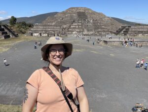 Veronica stands in front of a pyramid wearing a brown hat and glasses. She is smiling at the camera.