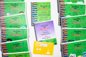 The components of the Building On Patterns Kindergarten, Second Edition Print Teacher's Kit on a white background. On either side are the green teacher's manuals laid out in lines, and in the middle are the teacher's booklet, the assessment booklet, and a stencil.