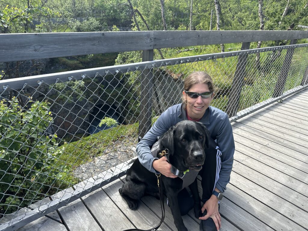 Hannah smiles, kneeling next to her guide dog who sits beside her with green trees and foliage in the background.