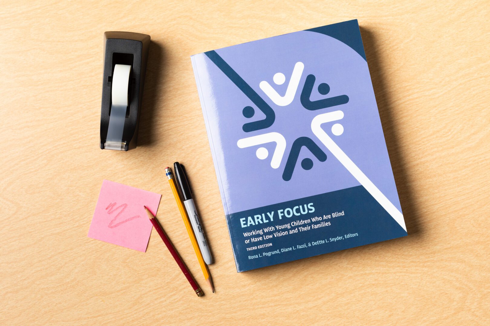 The blue Early Focus book on a wooden table next to a variety of writing instruments and a pink post it.