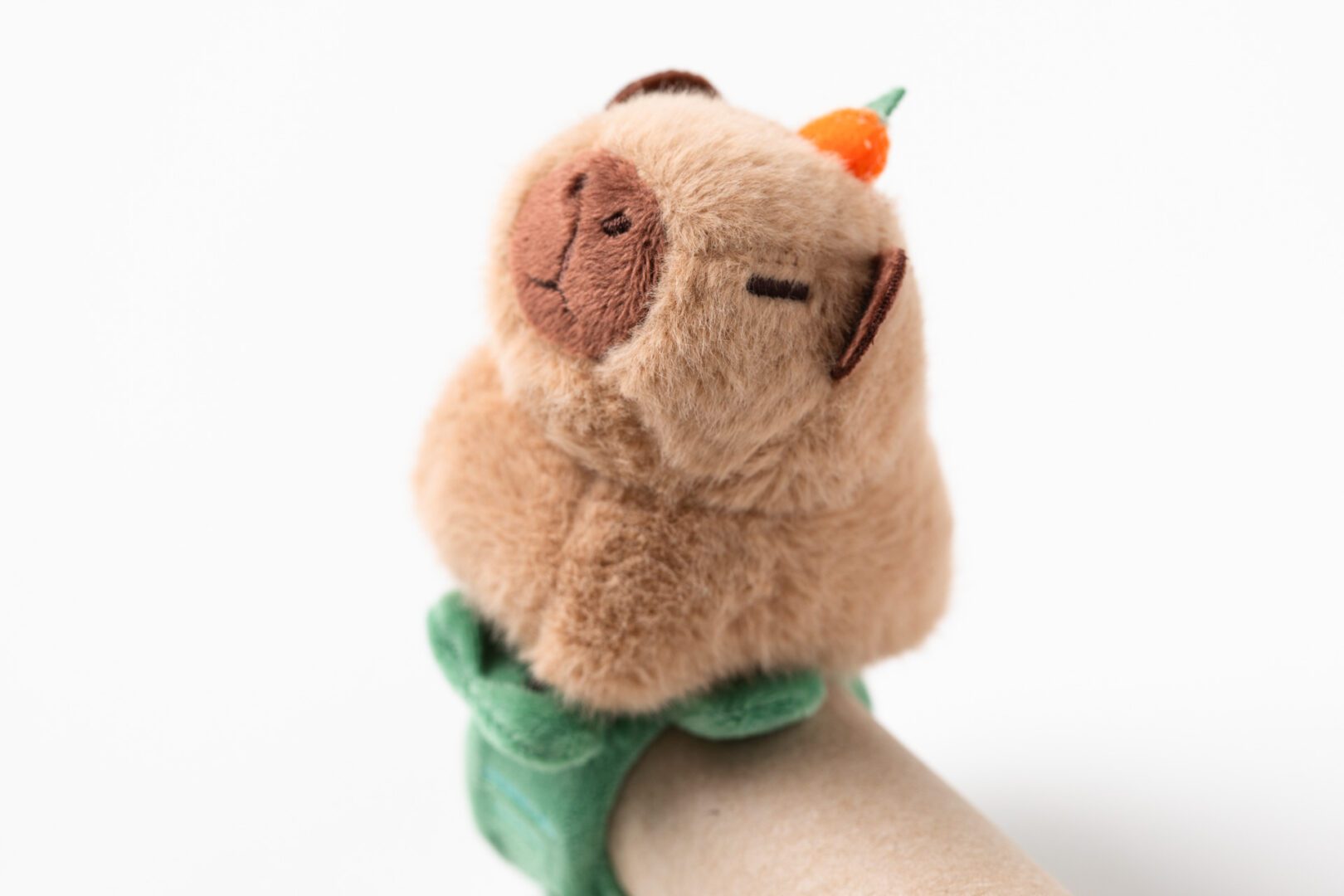 A plush capybara stuffie with a carrot on its head that sits at the top of a green snap bracelet.