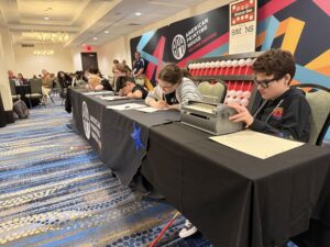 Students with focused expressions are seated at a long table with Cranmer abacuses, braillers, papers, and writing utensils on it. Behind them is a gigantic abacus and a backdrop with the APH logo on it.